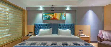 Une chambre d’hôtel avec un grand lit, une tête de lit capitonnée, un tableau au-dessus du lit, une table de chevet avec un téléphone et une fenêtre avec des stores.