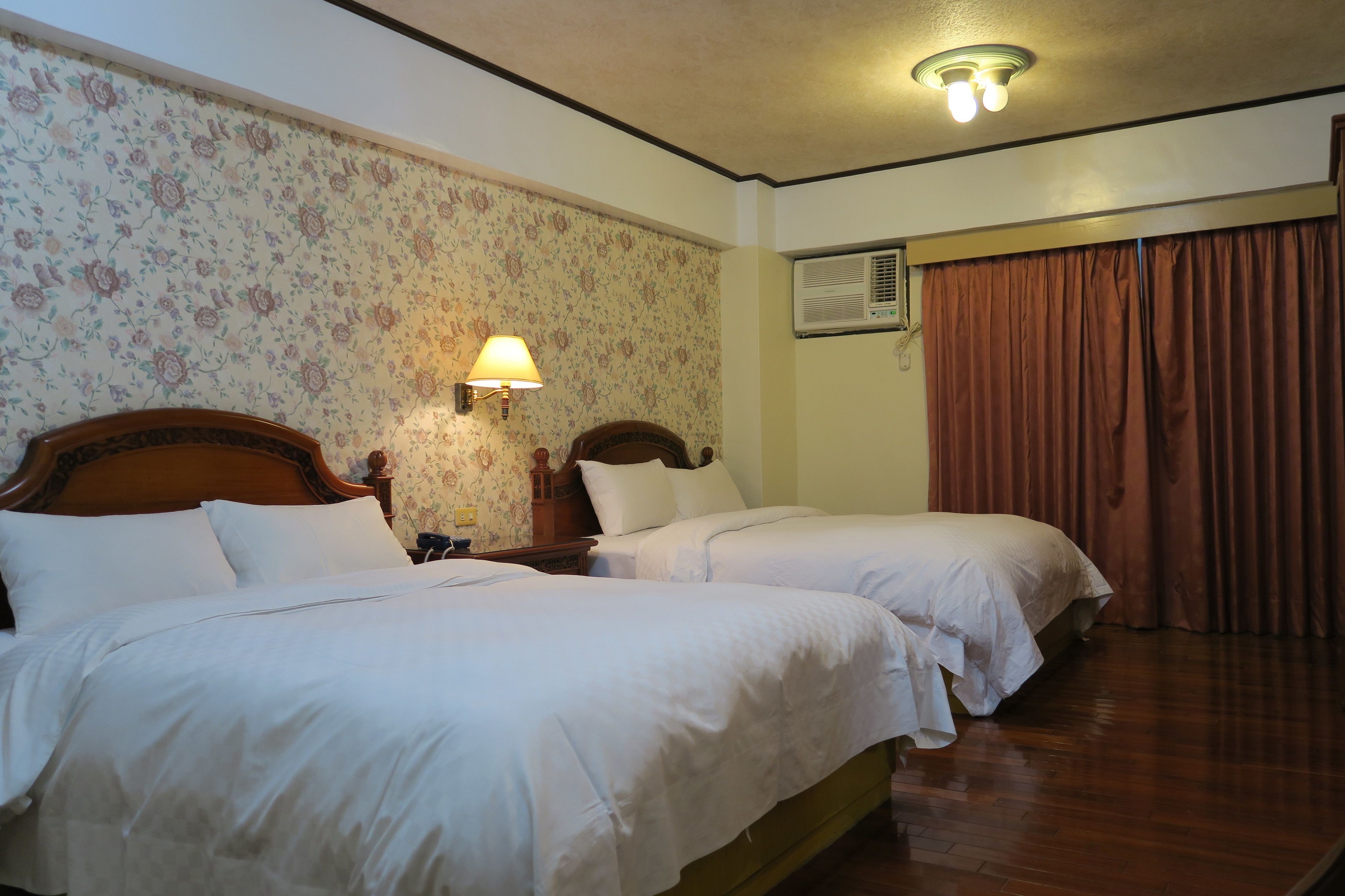 A hotel room with two beds, floral wallpaper, a wall-mounted air conditioner, and a ceiling light fixture.