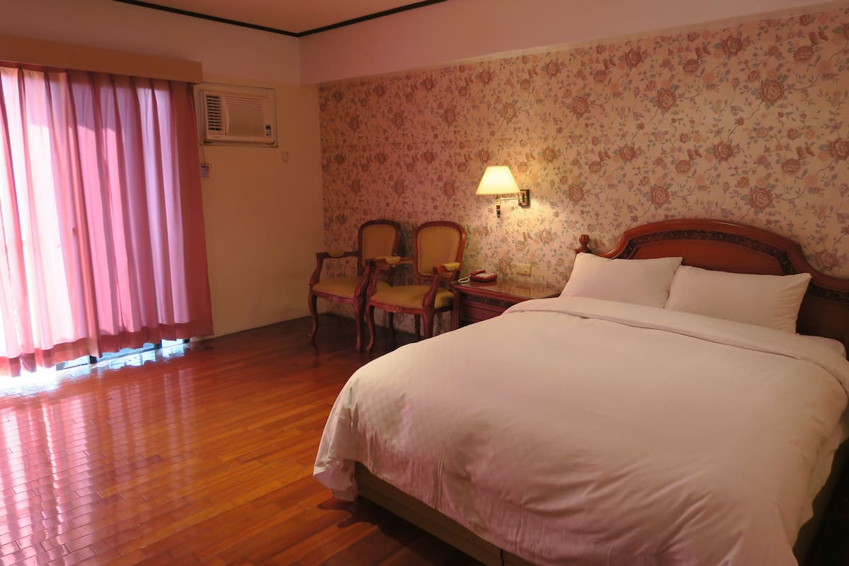 A bedroom with a wooden floor, a bed with white bedding, two wooden chairs, a nightstand, a wall-mounted lamp, and a floral wallpaper.