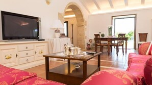 Een woonkamer met een televisie, een bijzettafeltje met decoratieve objecten, een bank en een eethoek met stoelen en een tafel.