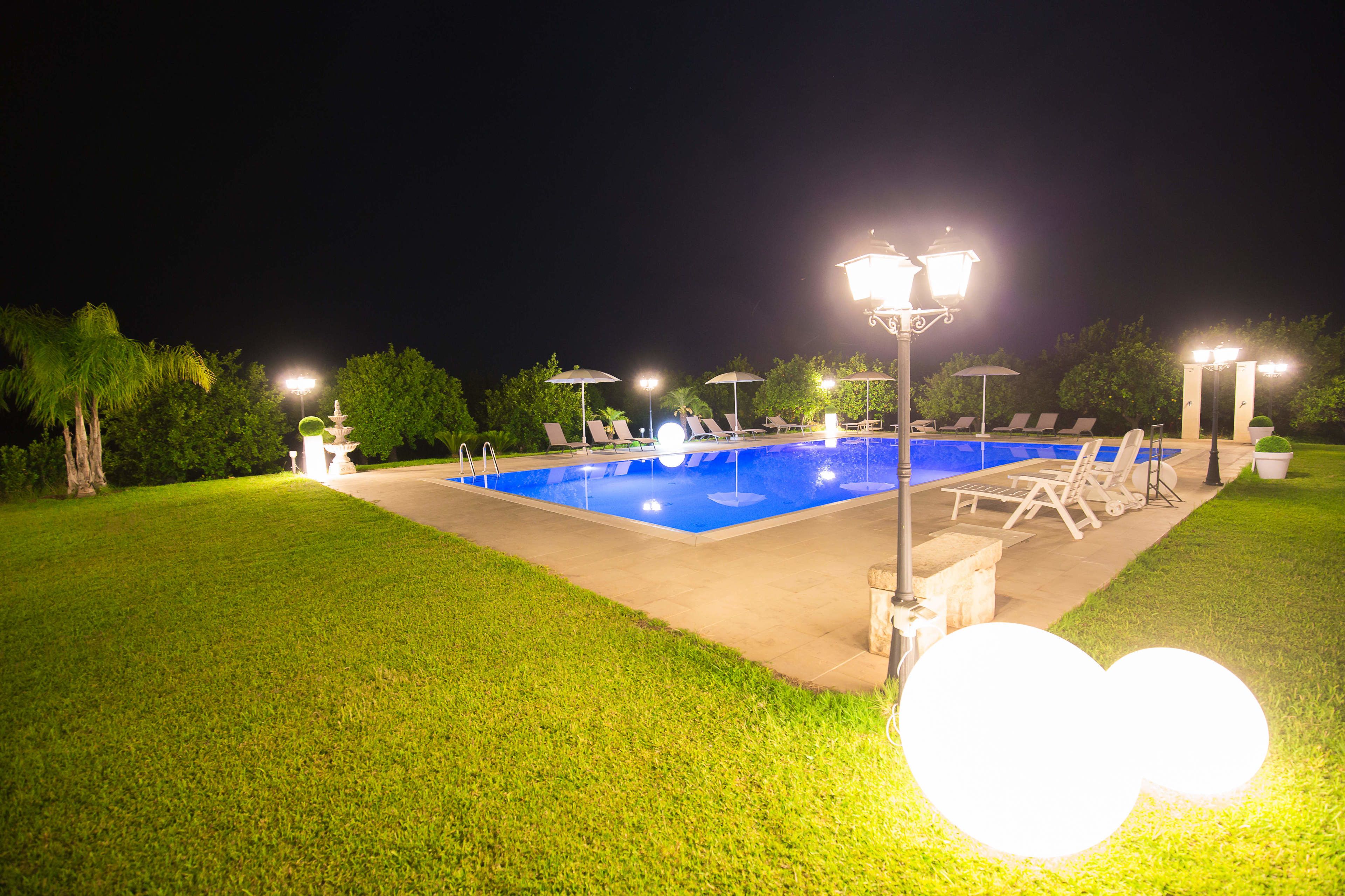 A nighttime swimming pool area with lounge chairs and umbrellas, surrounded by well-maintained grass and trees.