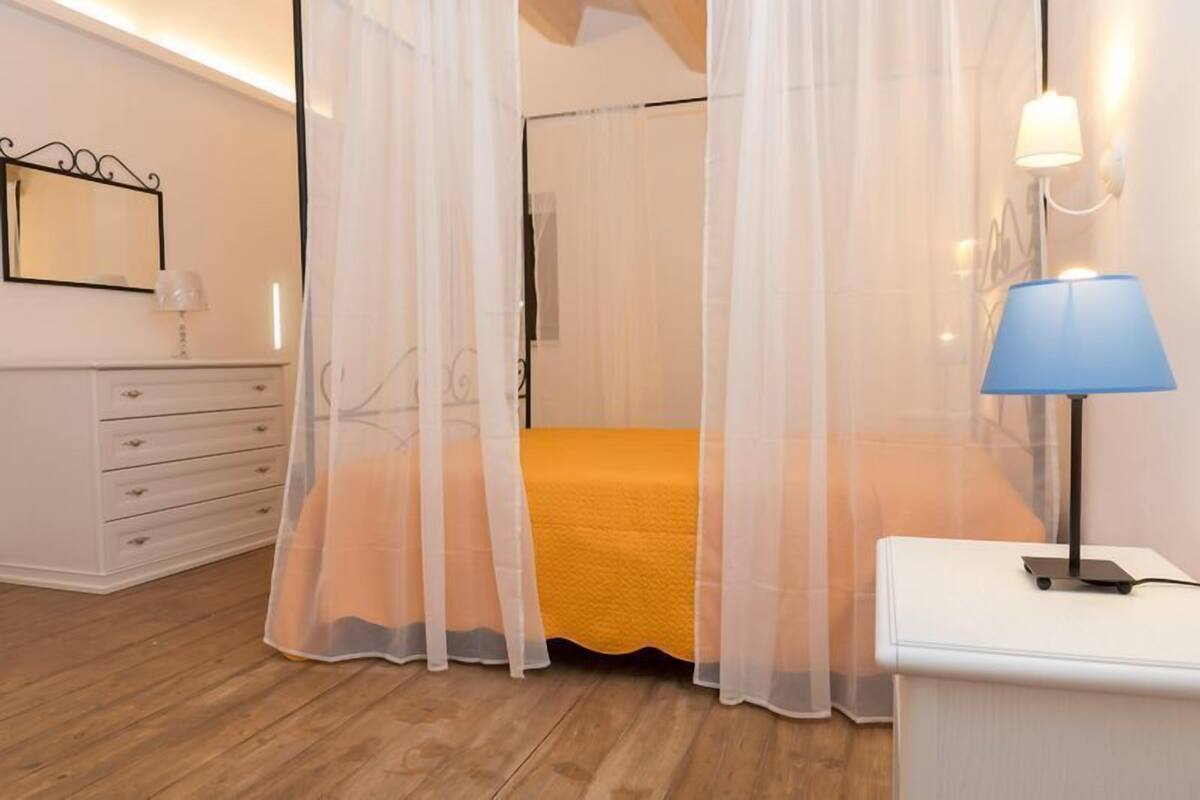 Ein Schlafzimmer mit einem Bett, das mit einem leichten Baldachin verziert ist, einem Nachttisch mit Lampe sowie einer Kommode mit Schubladen.