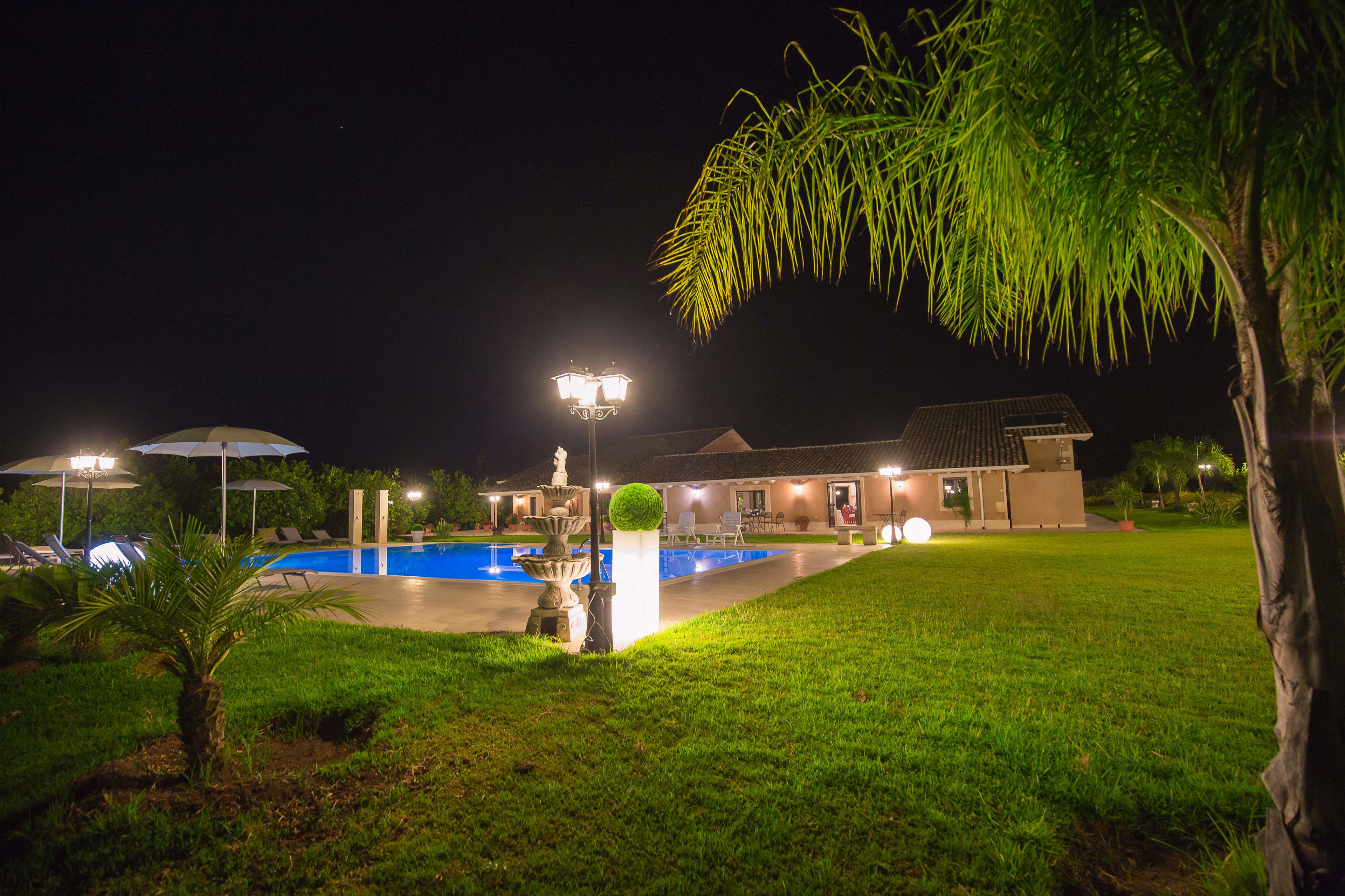 Vue nocturne d’un espace piscine avec une fontaine, des parasols et un bâtiment doté d’un toit recouvert de tuiles.