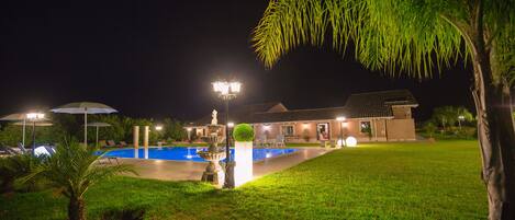 Vue nocturne d’un espace piscine avec une fontaine, des parasols et un bâtiment doté d’un toit recouvert de tuiles.