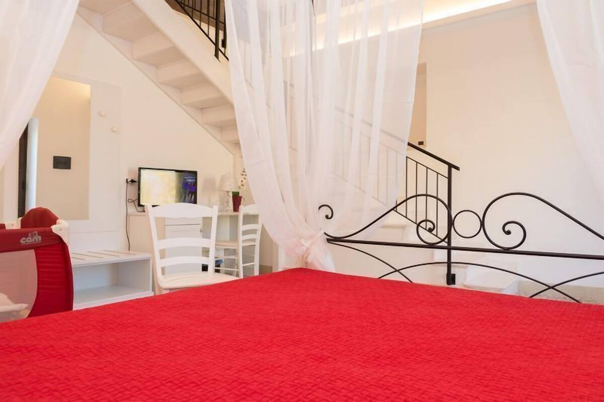 Ein Schlafzimmer mit roter Bettdecke, weißen Vorhängen und einer Treppe im Hintergrund.