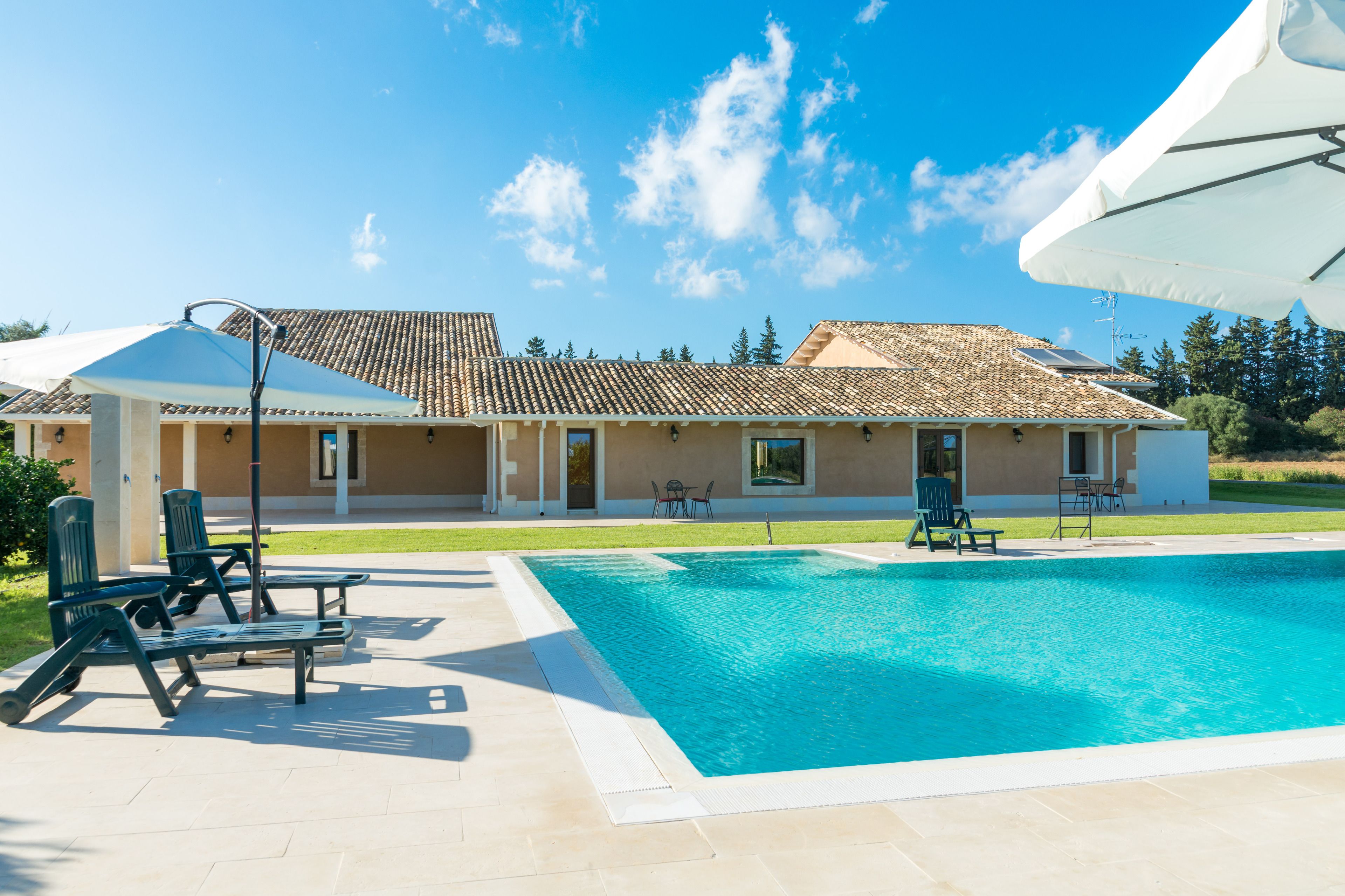 A swimming pool with lounge chairs and umbrellas, a building with a tiled roof, and a clear blue sky.