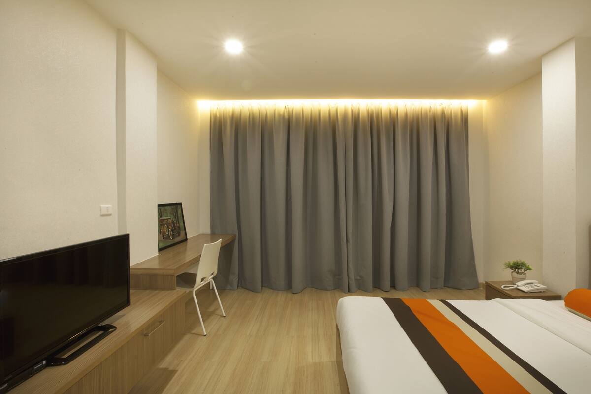 A modern hotel room with a flat-screen TV, a desk, a bed with an orange and white striped bedspread, and a large window with grey curtains.