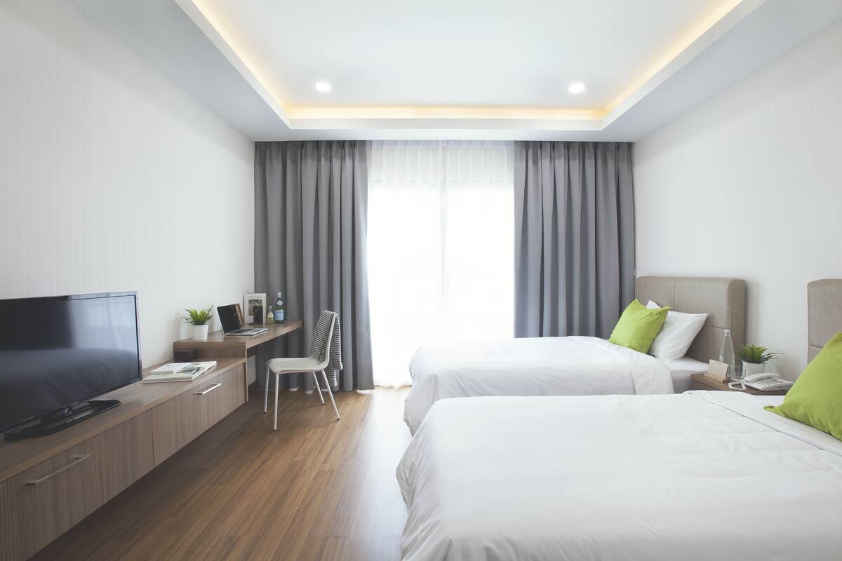A modern hotel room with a flat-screen TV, a desk with a laptop, two beds with white bedding and green pillows, a chair, and a bedside table with a lamp and a phone.
