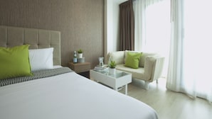 Quarto de hotel moderno com uma cama grande, um sofá, uma mesinha com uma planta e uma mesa de cabeceira com livros e uma luminária.