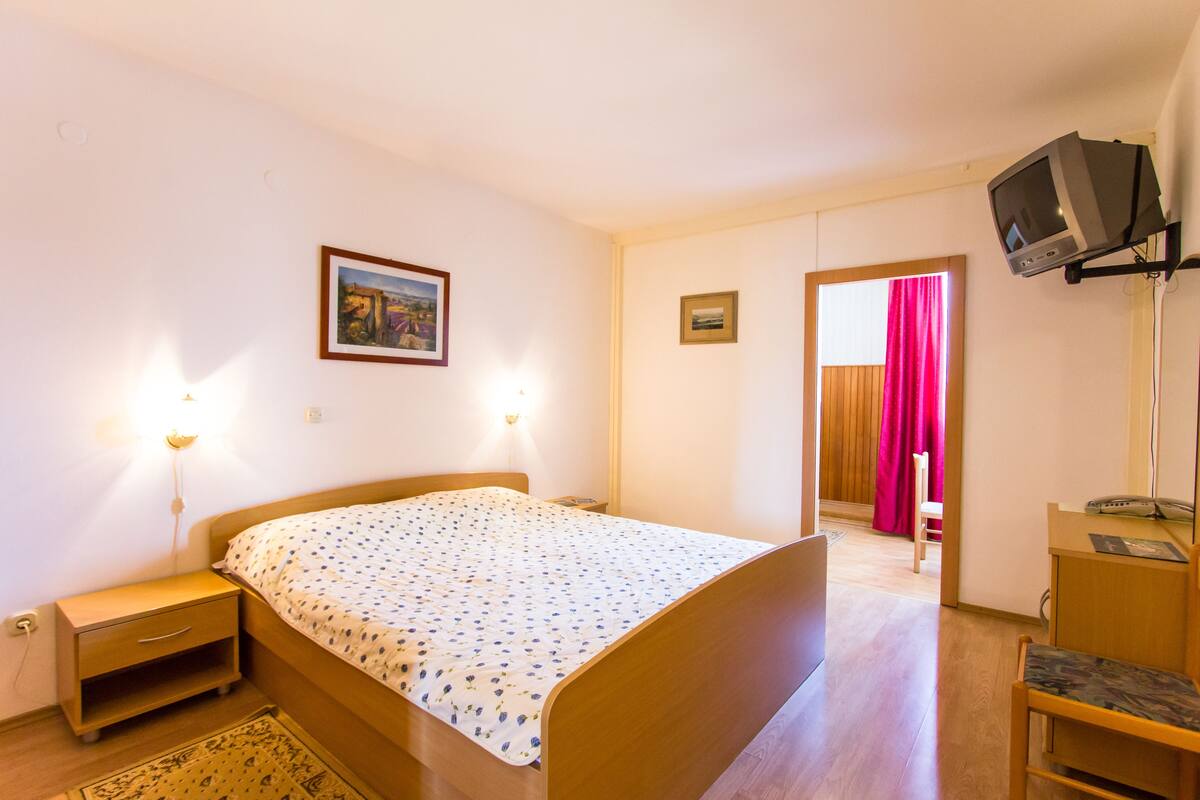A bedroom with a wooden bed, bedside lamps, a small desk, a chair, a television mounted on the wall, and a door leading to another room.