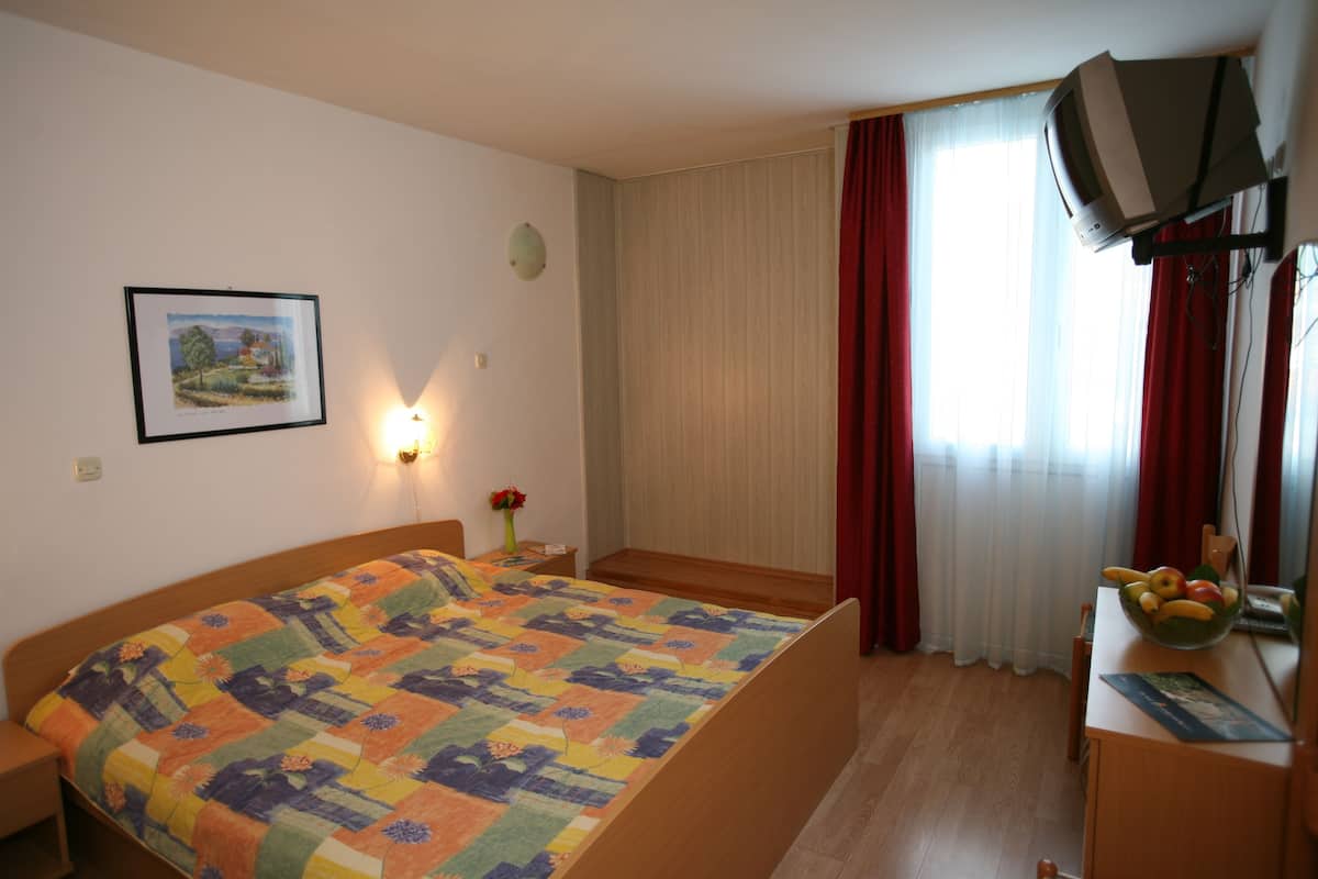 A hotel room with a bed, a desk with a fruit bowl, a television, a painting on the wall, and a window with curtains.