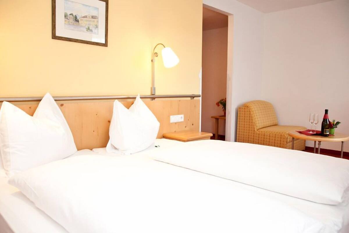 Habitación de hotel con una cama, una silla, una mesita con una botella y vasos, y una lámpara.