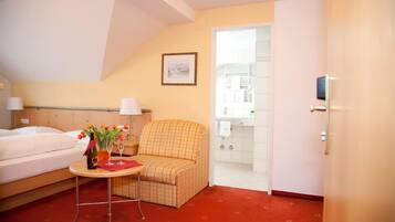 A hotel room with a bed, a small table with a vase of flowers, a patterned armchair, and a view of the bathroom.