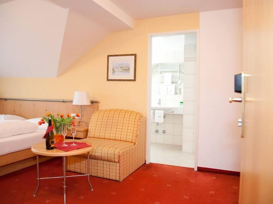 A hotel room with a bed, a small table with a vase of flowers, a patterned armchair, and a view of the bathroom.