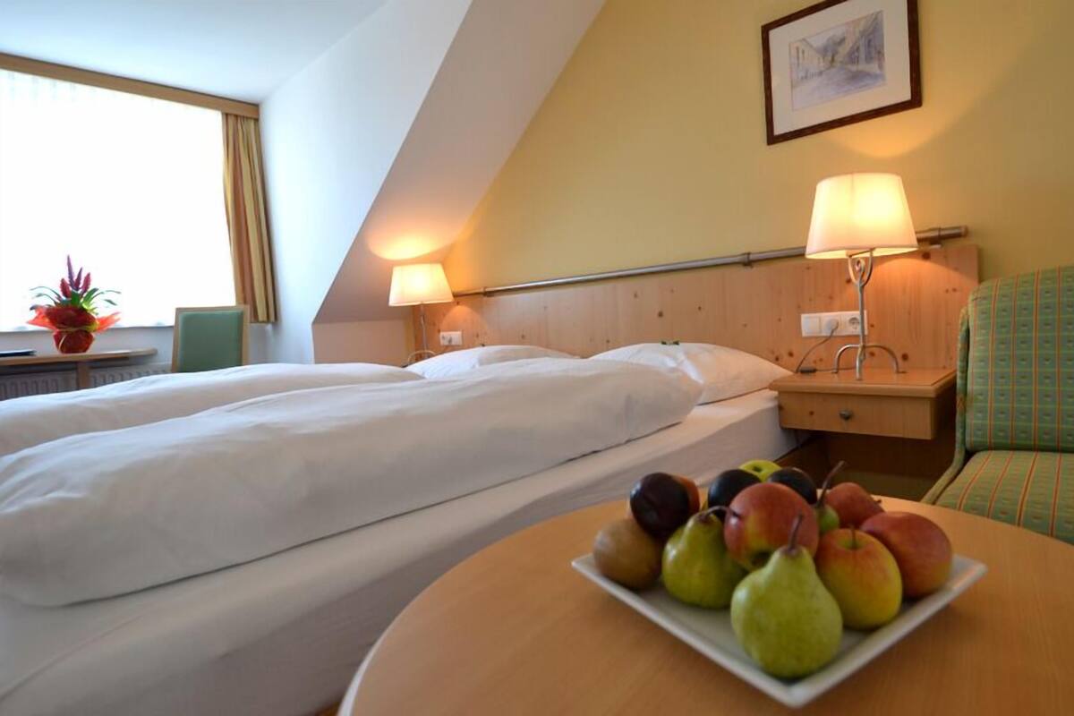 Habitación de hotel con dos camas, una mesita de noche con lámpara, una pequeña mesa con fruta y una ventana con cortinas.