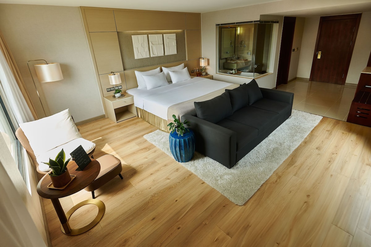 Quarto de hotel moderno com uma cama grande, um sofá, uma mesinha com uma planta e uma mesa de cabeceira com abajur.