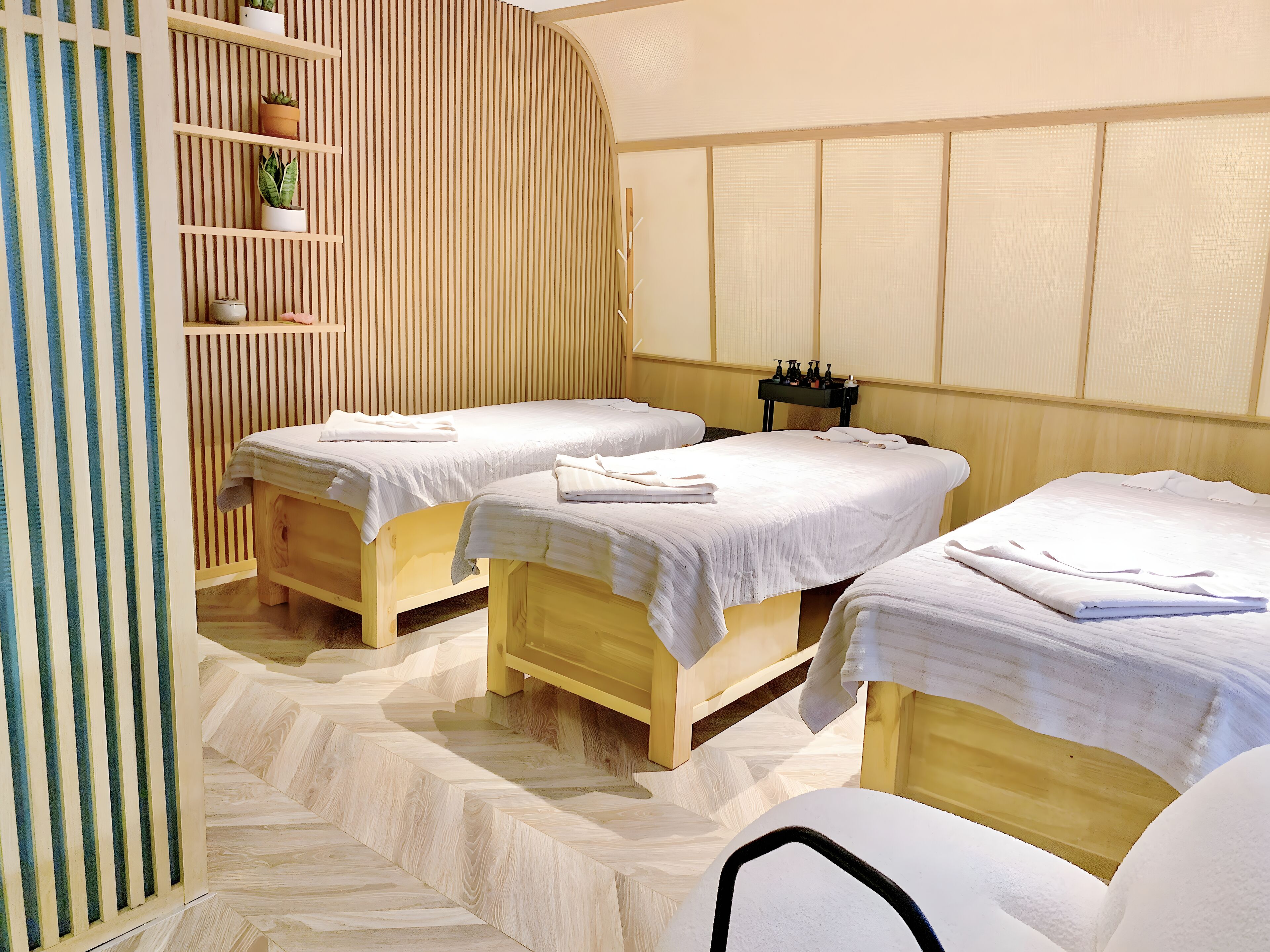 A room with three wooden beds, each with white linens and towels, a shelf with potted plants, and a wooden slatted wall.