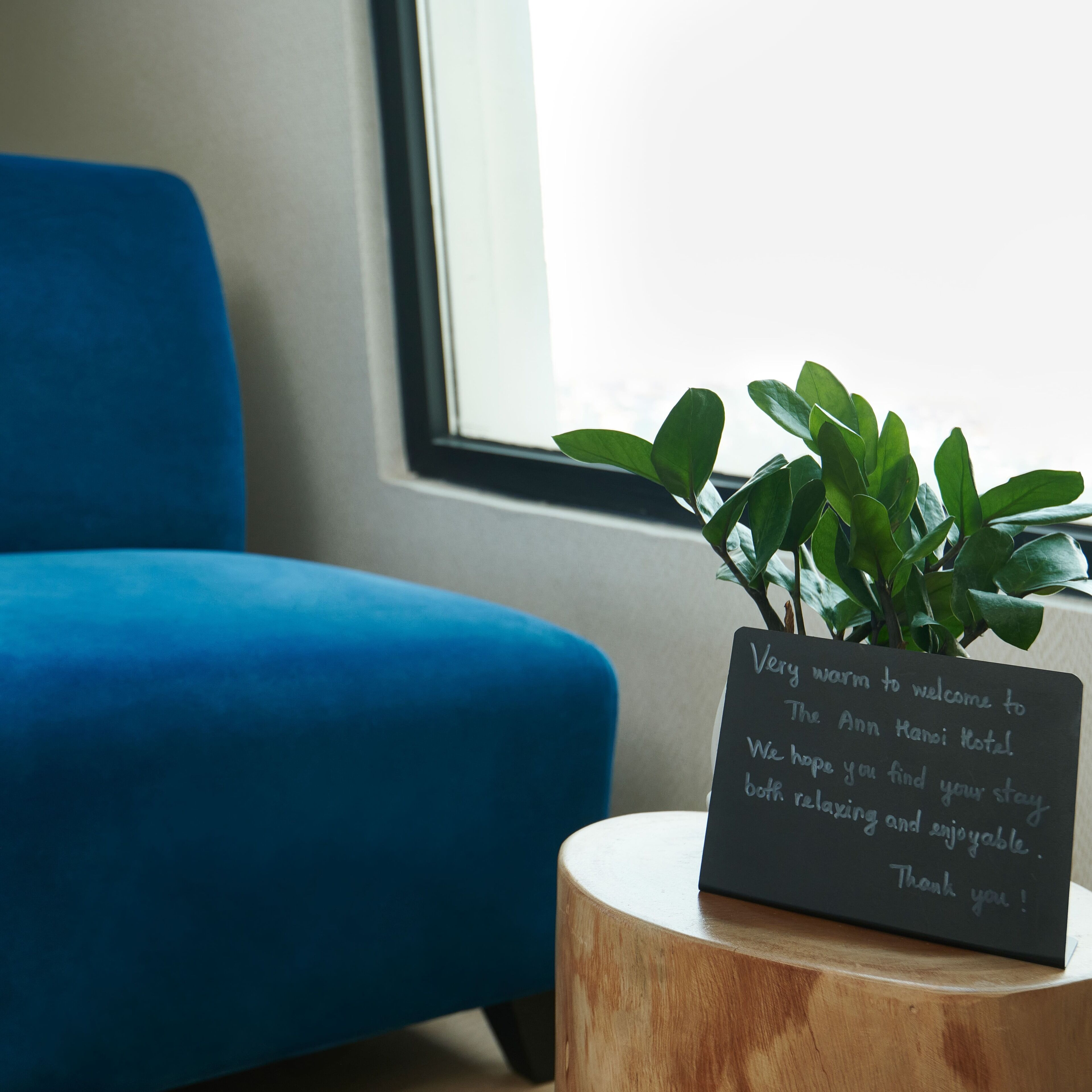 Un letrero en un taburete de madera con un mensaje dando la bienvenida a los huéspedes al Hotel The Ann Hanoi, ubicado frente a una silla azul y una ventana.