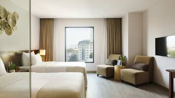 Ein Hotelzimmer mit zwei Betten, einem Sofa, einem Sessel, einem Fernseher und einem großen Fenster mit Blick auf die Stadt.