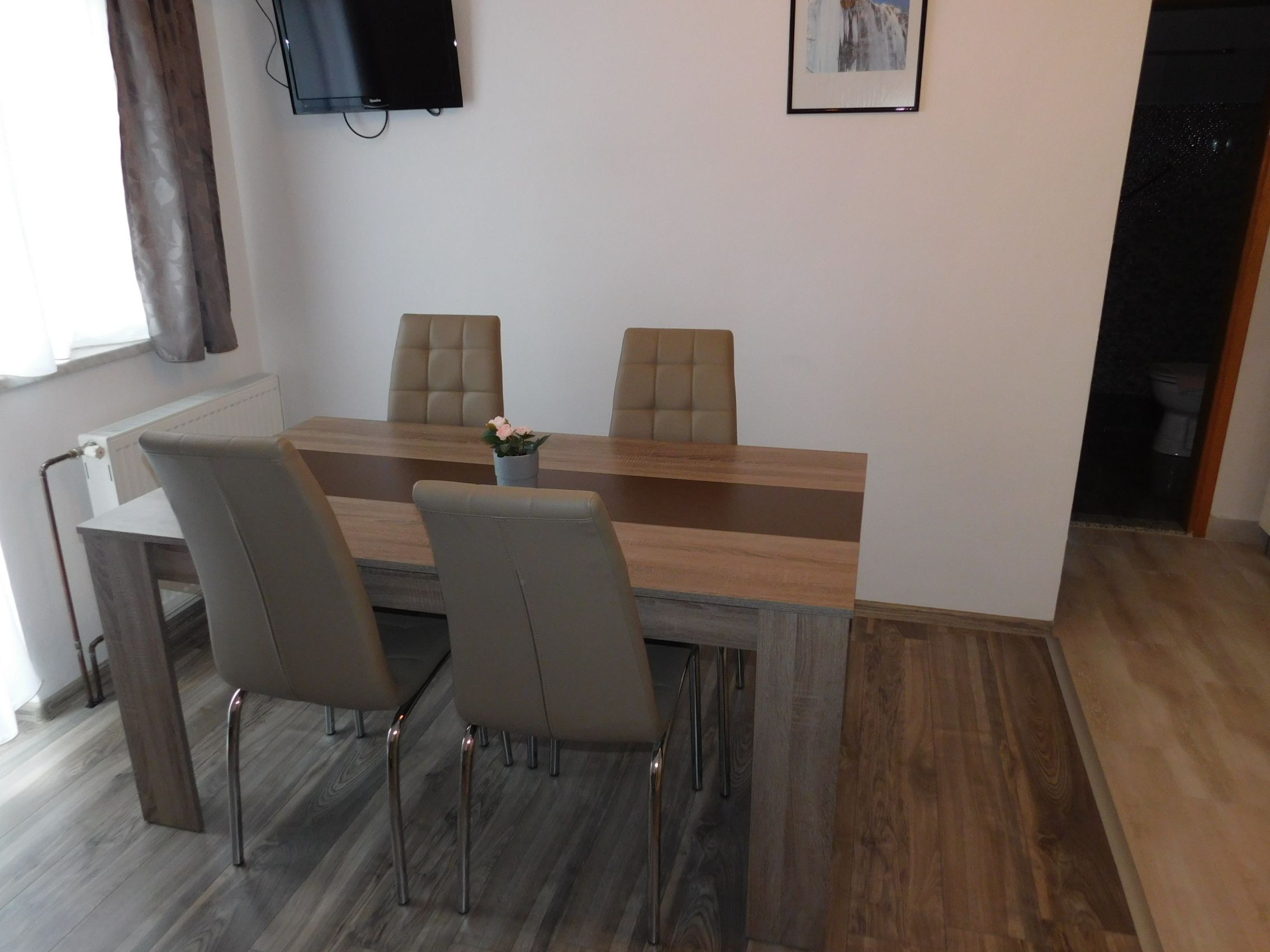 Un comedor moderno con una mesa de madera y cuatro sillas, un televisor montado en la pared y un cuadro enmarcado sobre la mesa.