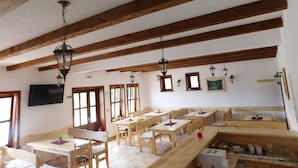 Ein Essbereich mit Holztischen und -bänken, einem an der Wand befestigten Fernseher und einer Decke mit freiliegenden Holzbalken.