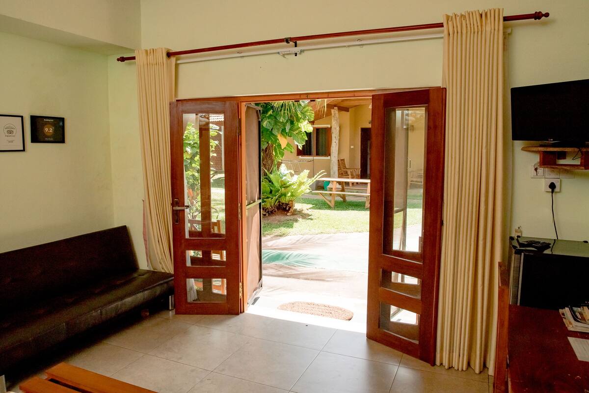 Sala de estar com sofá, televisão e porta que dá para área externa com rede e vegetação.
