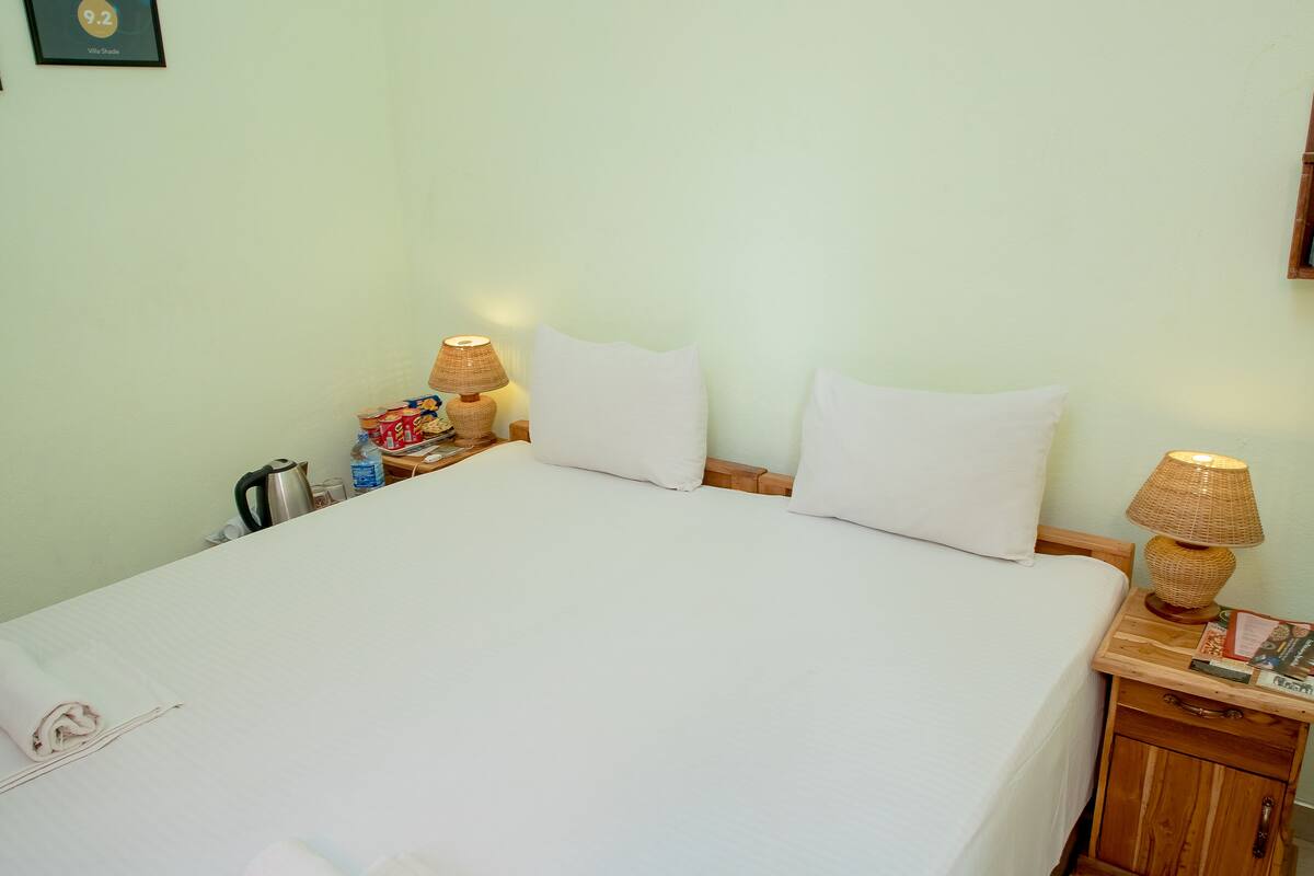 Cama bem arrumada com lençóis brancos, duas luminárias de cabeceira e uma mesa de cabeceira de madeira com uma chaleira e uma garrafa.
