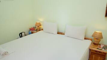 Una cama bien hecha con sábanas blancas, dos lámparas de noche y una mesita de madera con una tetera y una botella.