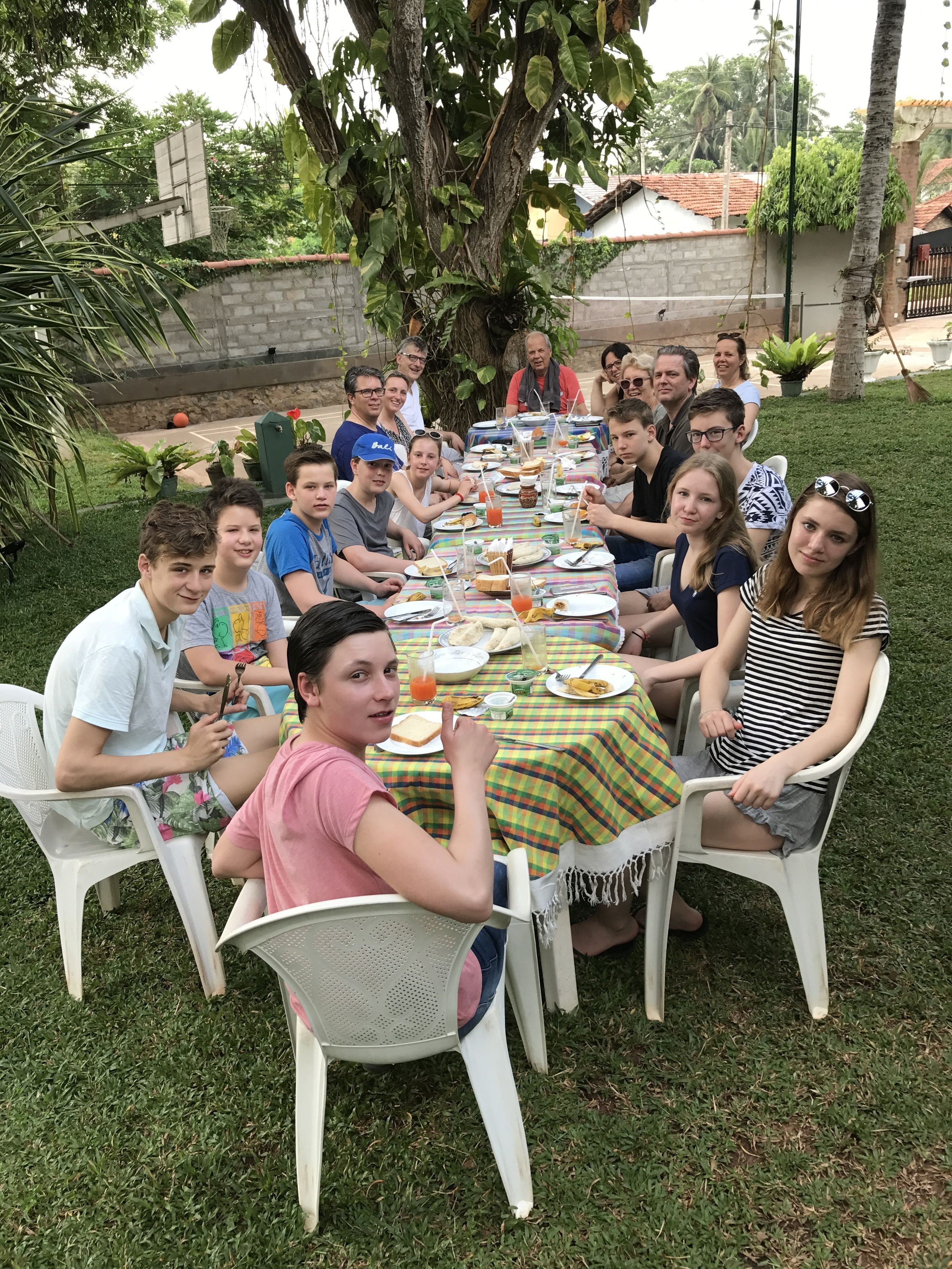 Un grupo de personas sentadas en una mesa al aire libre con mantel a cuadros, rodeado de follaje y un árbol.