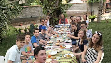 Un grupo de personas sentadas en una mesa al aire libre con mantel a cuadros, rodeado de follaje y un árbol.