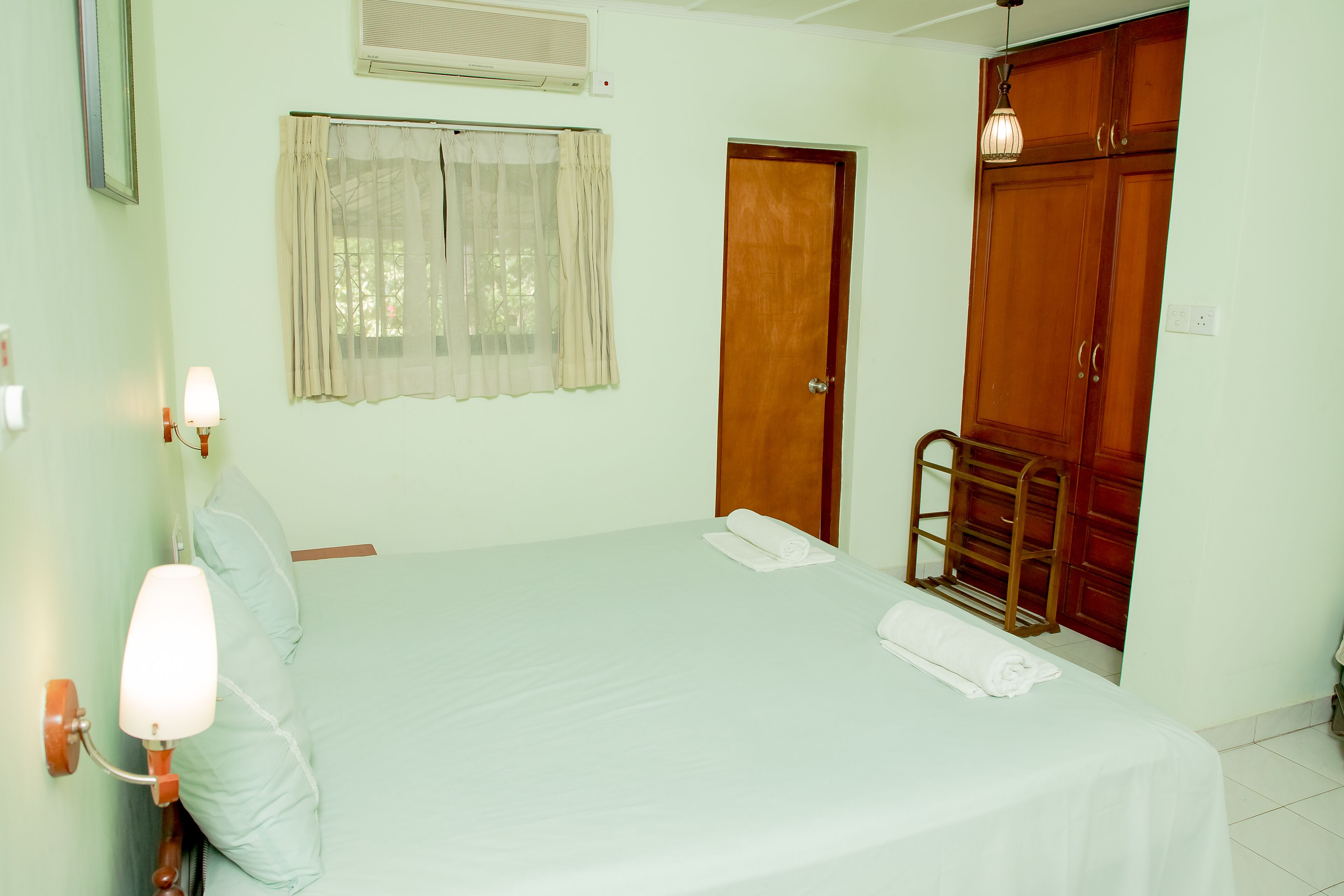 Un dormitorio con cama, mesita de noche, lámpara de pared, ventana con cortinas, armario de madera y puerta.