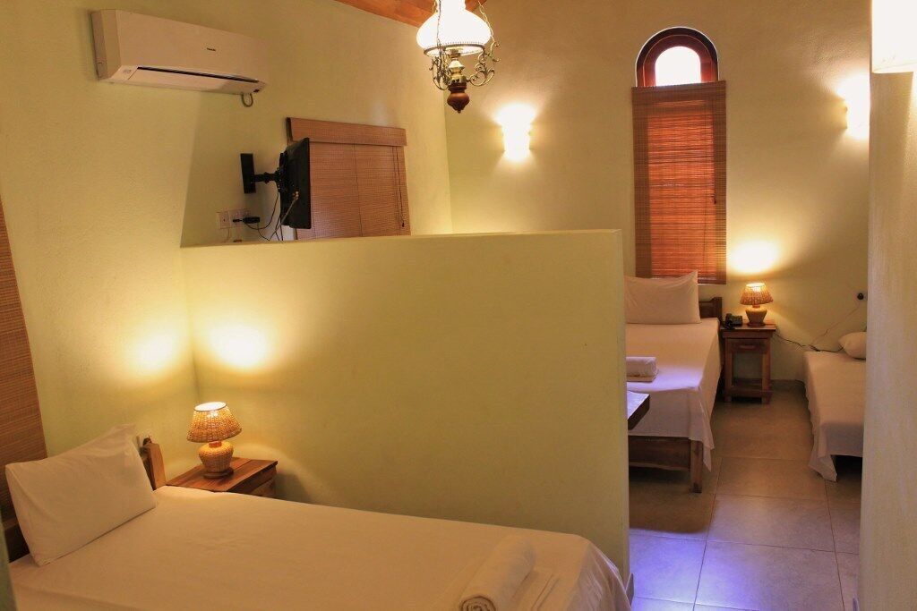 A hotel room with a bed, bedside lamp, and a small table. There is a mirror on the wall and a window with blinds.
