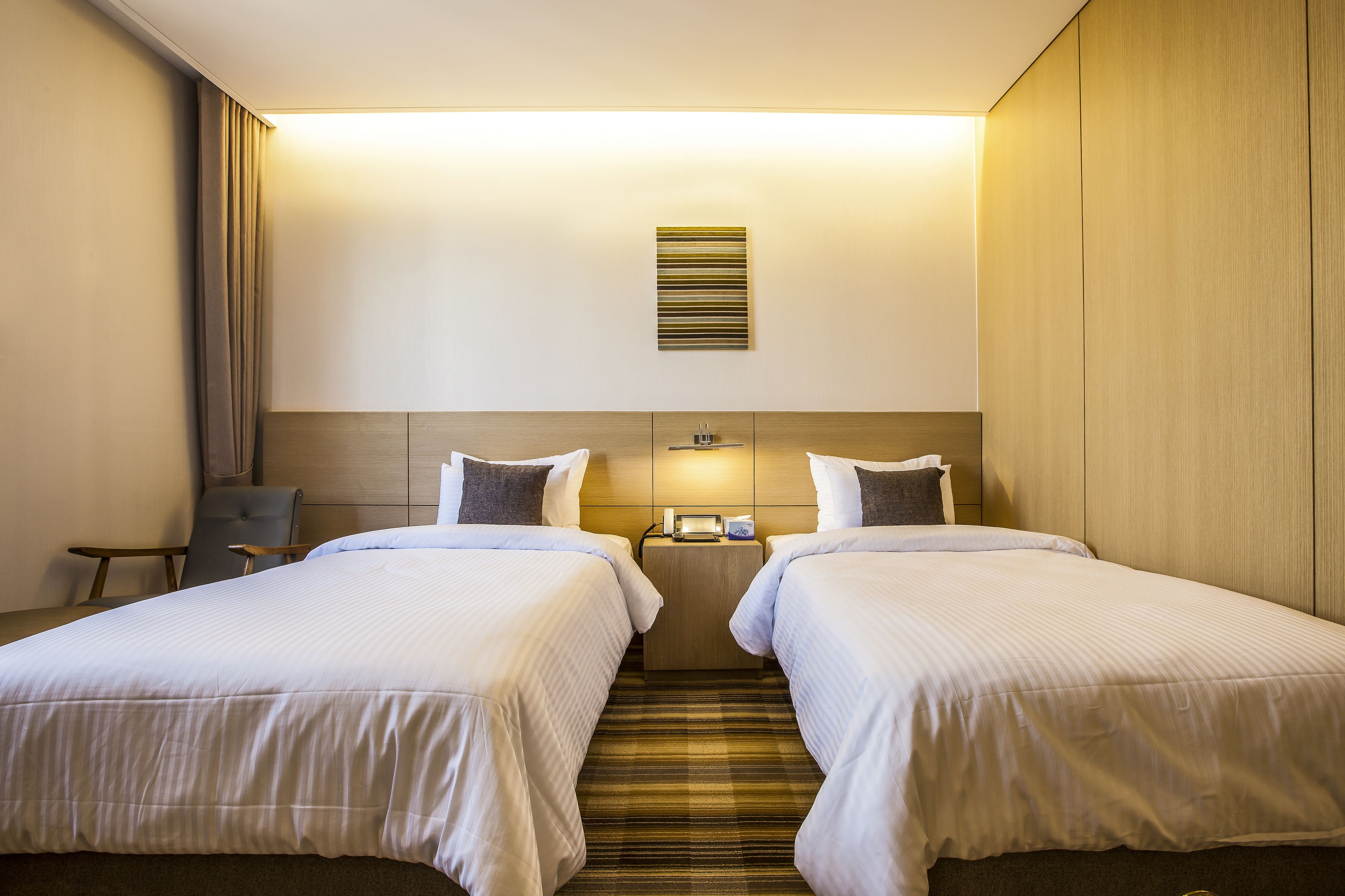 A hotel room with two beds, a small table, a chair, a wall-mounted light, and a striped carpet.