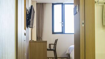 A hotel room with a bed, a desk, a chair, a television mounted on the wall, and a window with curtains.