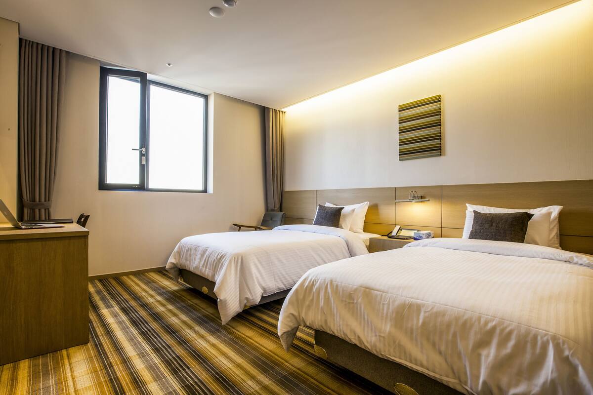 A hotel room with two beds, a desk, a chair, a window, and a wall-mounted artwork.