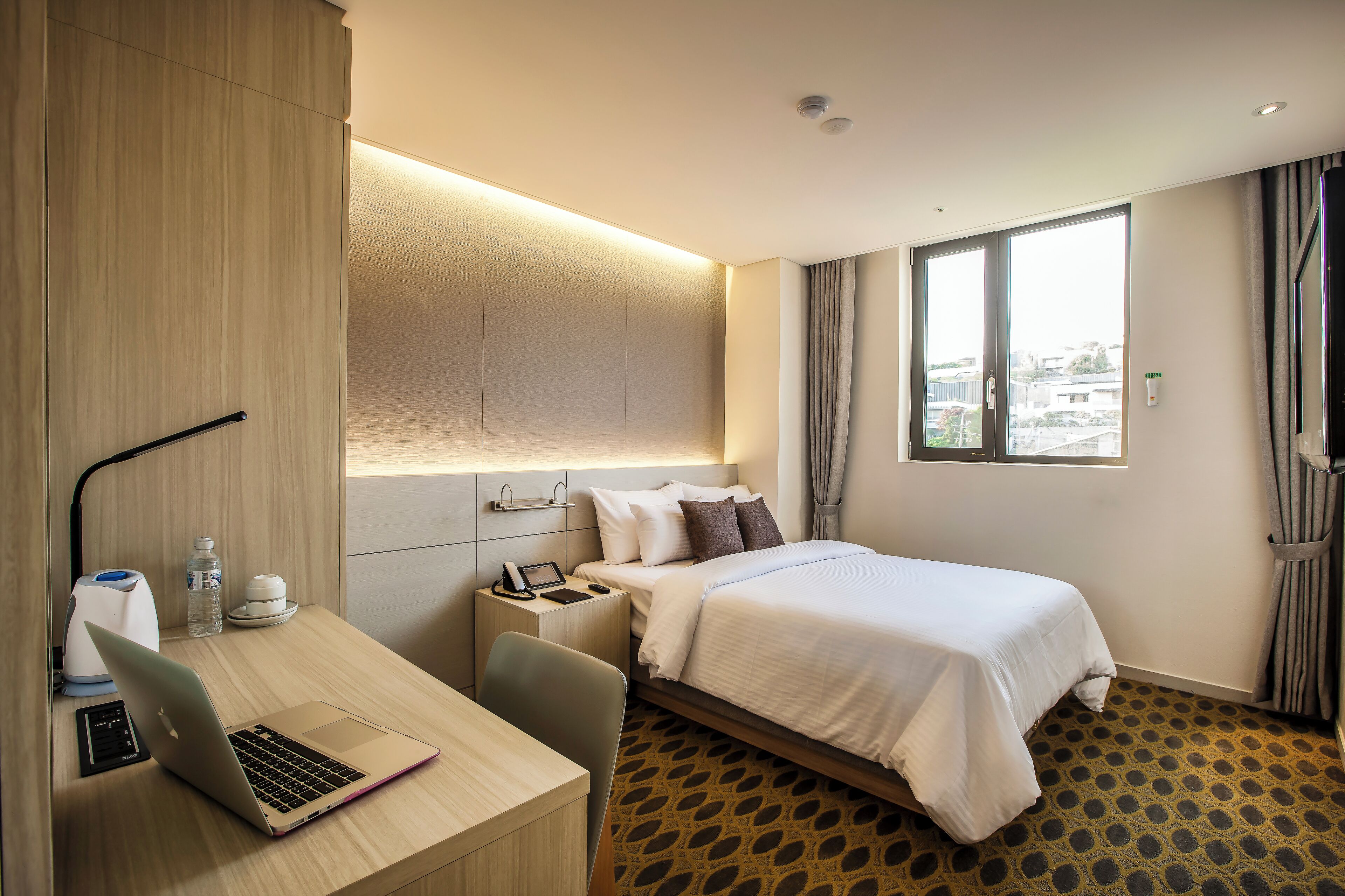 Une chambre d’hôtel avec un lit, un bureau avec un ordinateur portable, une table de chevet avec un téléphone et une bouteille, et une fenêtre avec des rideaux.