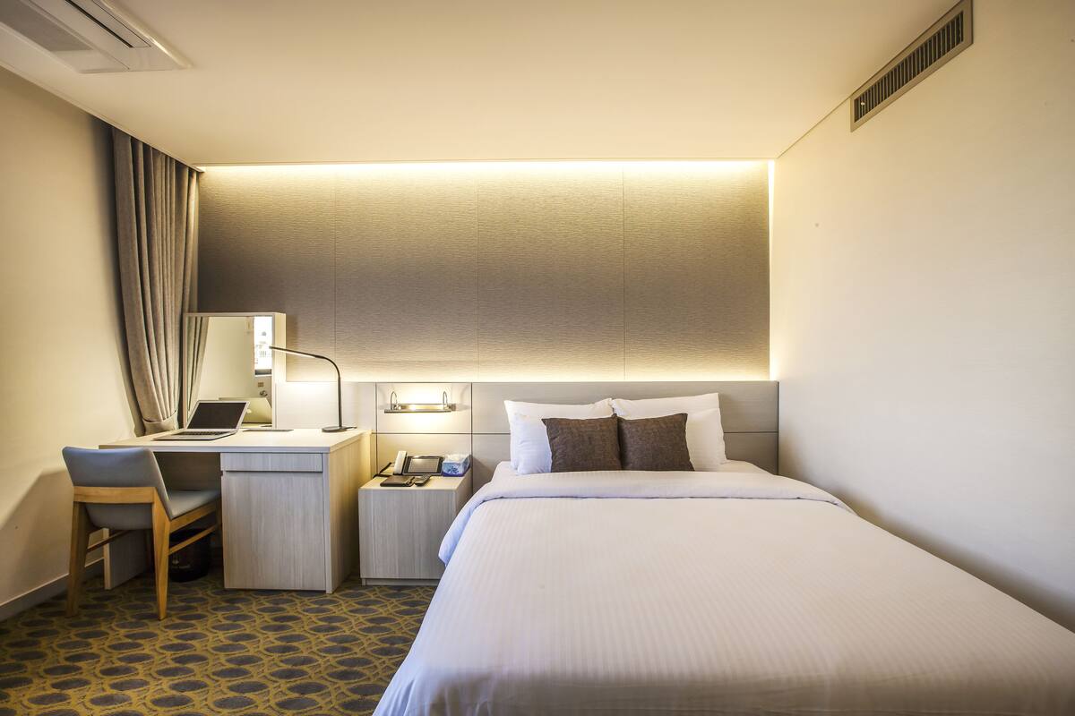 A hotel room with a bed, desk, chair, and a small table with a lamp.