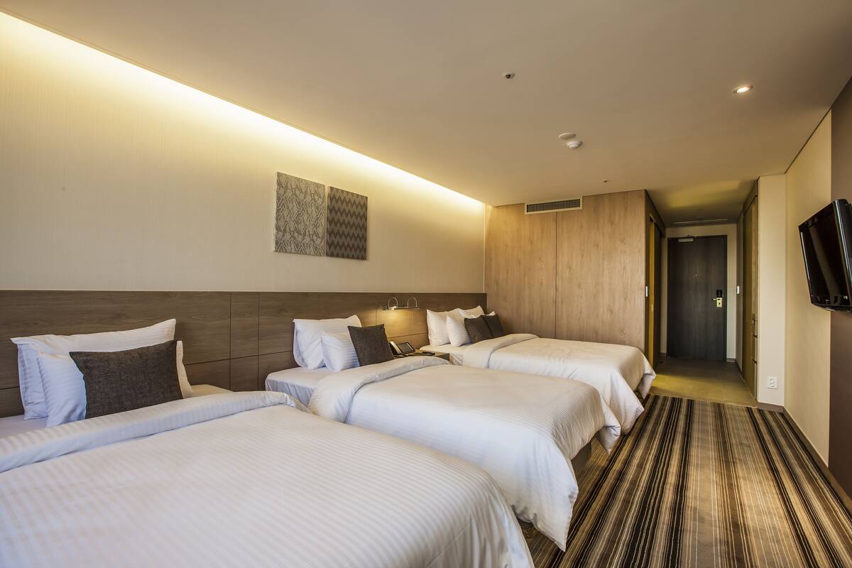 A hotel room with three beds, a wooden headboard, a patterned wall hanging, and a television on the wall.