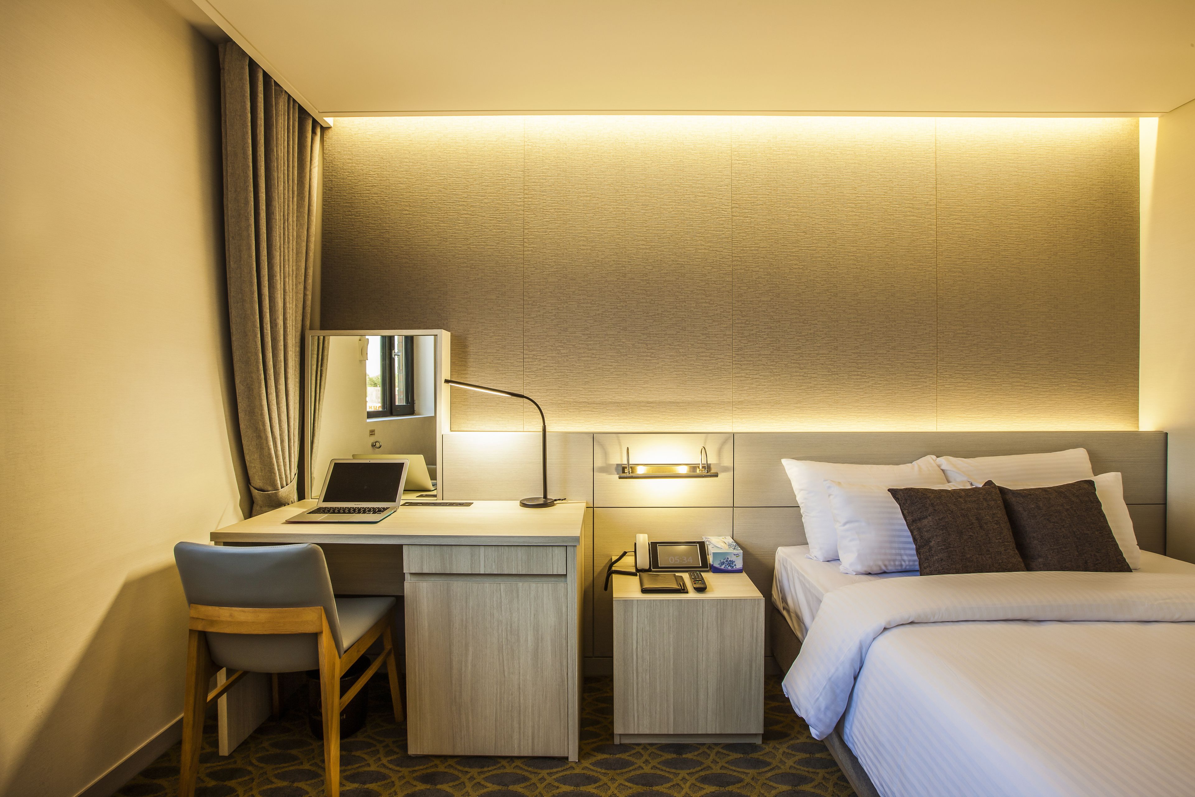 Habitació d'hotel amb un llit, una taula de treball amb un ordinador portàtil, una cadira, una taula de nit amb un telèfon i un comandament a distància, i una làmpada penjada a la paret.