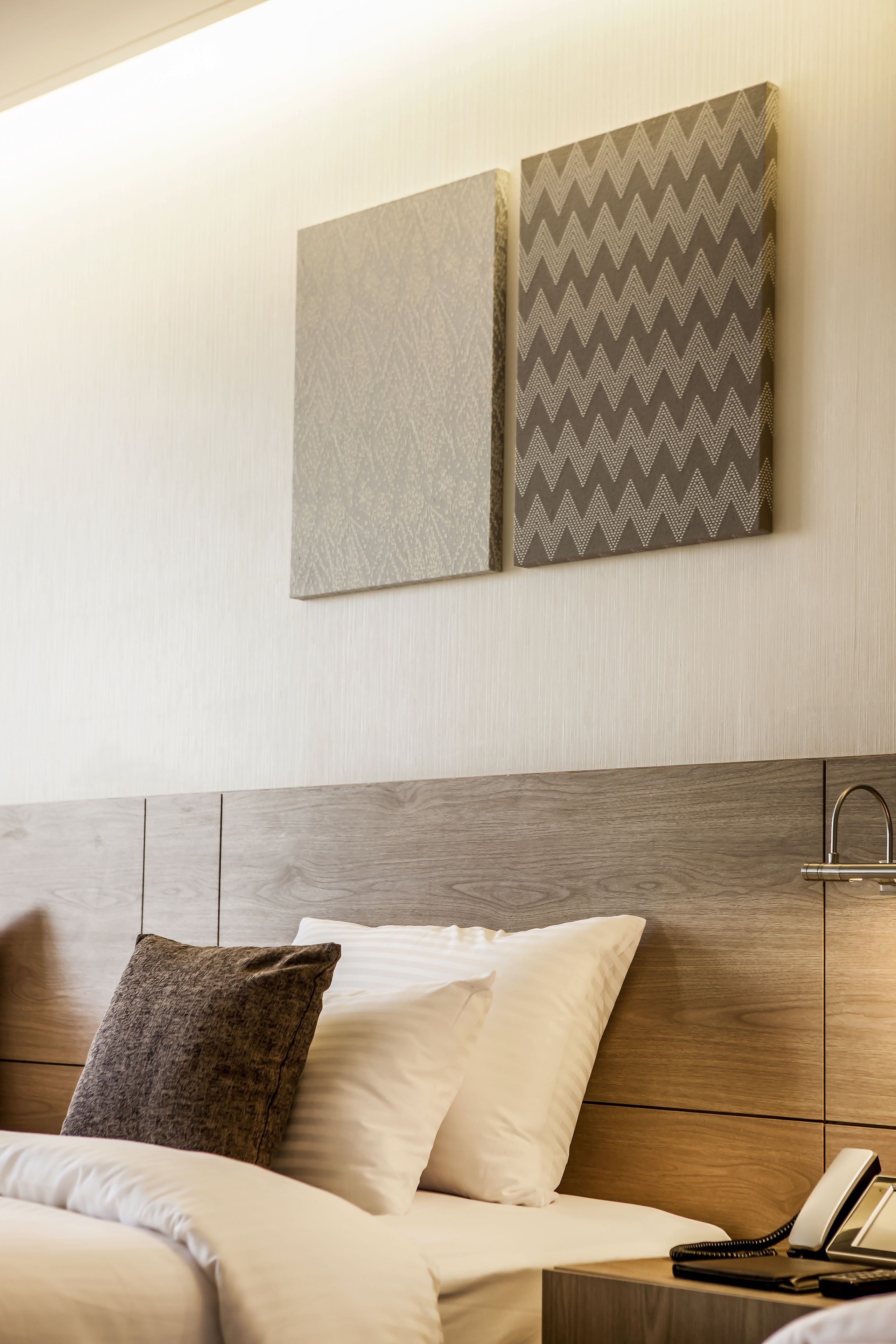 Habitación de hotel con cabecera de madera, cama con ropa blanca, dos almohadas, mesita de noche con teléfono y dos cuadros en la pared.