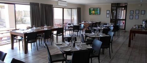 Un comedor amplio con varias mesas y sillas, ventanas grandes con cortinas y un televisor de pantalla plana montado en la pared.