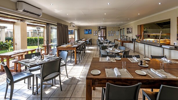 A spacious dining area with multiple tables and chairs, large windows offering views of the outdoor patio, and a kitchen area visible through the glass partition. - Kathu Inn by Country Hotels (Kathu)