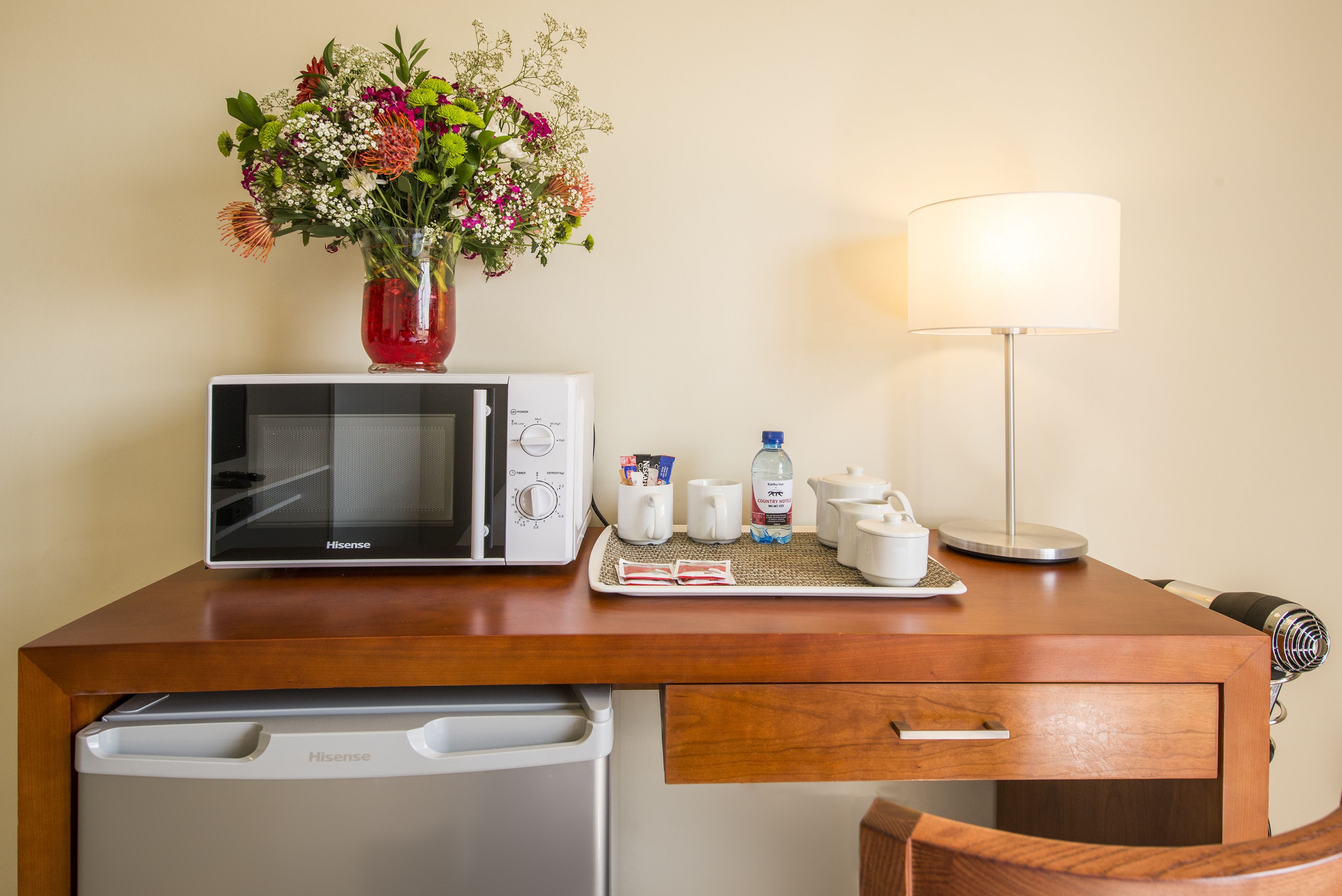 A hotel room with a microwave, a Hisense TV, a small table with a vase of flowers, a lamp, and a coffee maker.