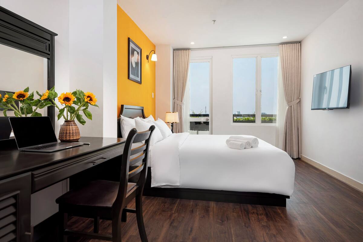 A hotel room with a bed, desk, laptop, and a vase of sunflowers.
