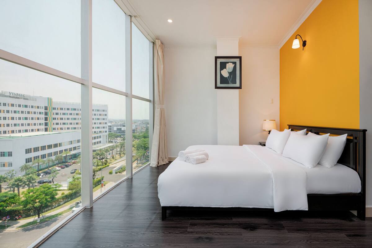 A hotel room with a large window, a bed with white linens, a nightstand with a lamp, and a framed picture on the wall.