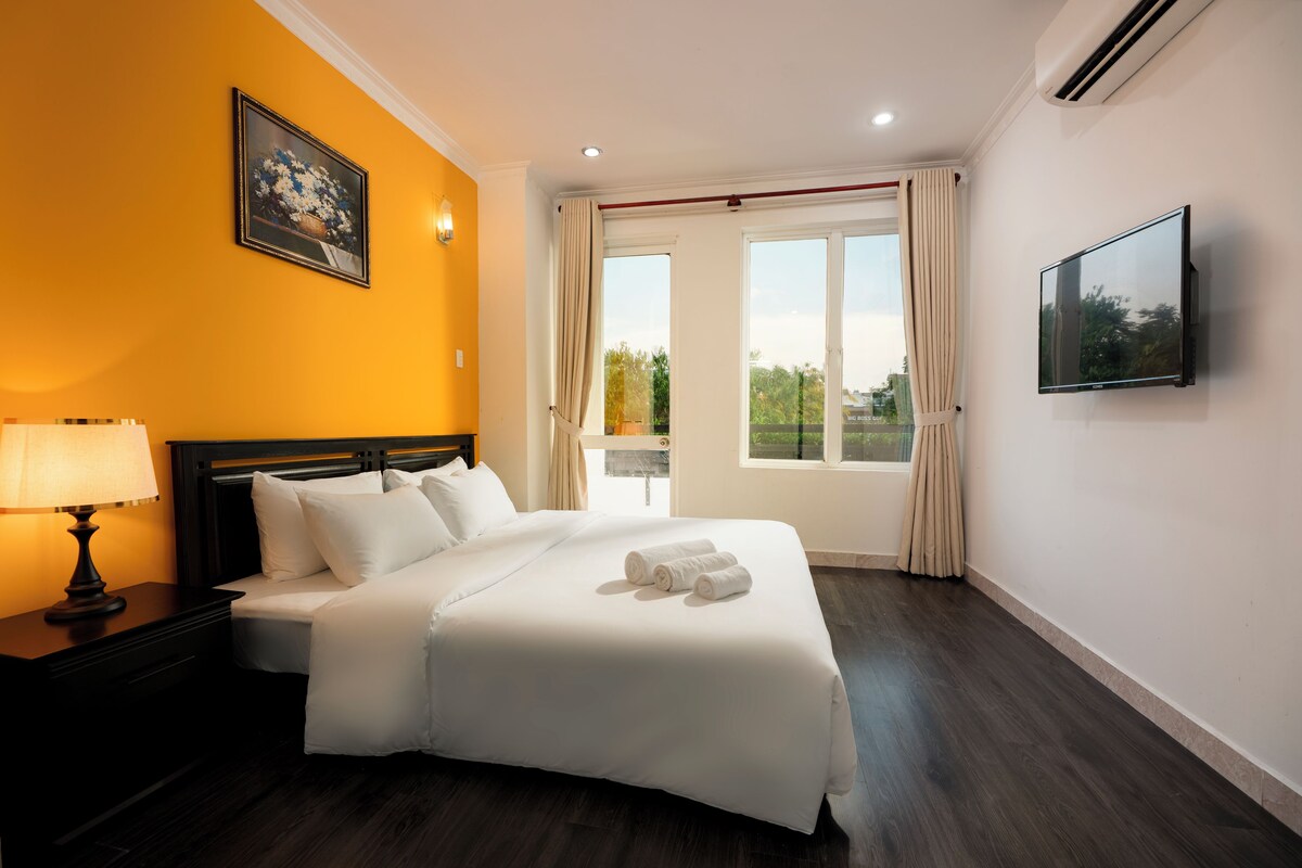 A hotel room with a large bed, a nightstand with a lamp, a flat-screen TV, and a window with curtains.