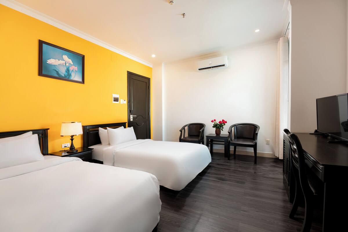 A hotel room with two beds, a desk, a TV, a chair, and a small table with flowers.