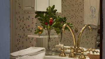 Une salle de bain avec un lavabo blanc, des robinets dorés et un miroir. Une serviette est accrochée au mur et un panier contenant des articles de toilette est posé sur le comptoir.