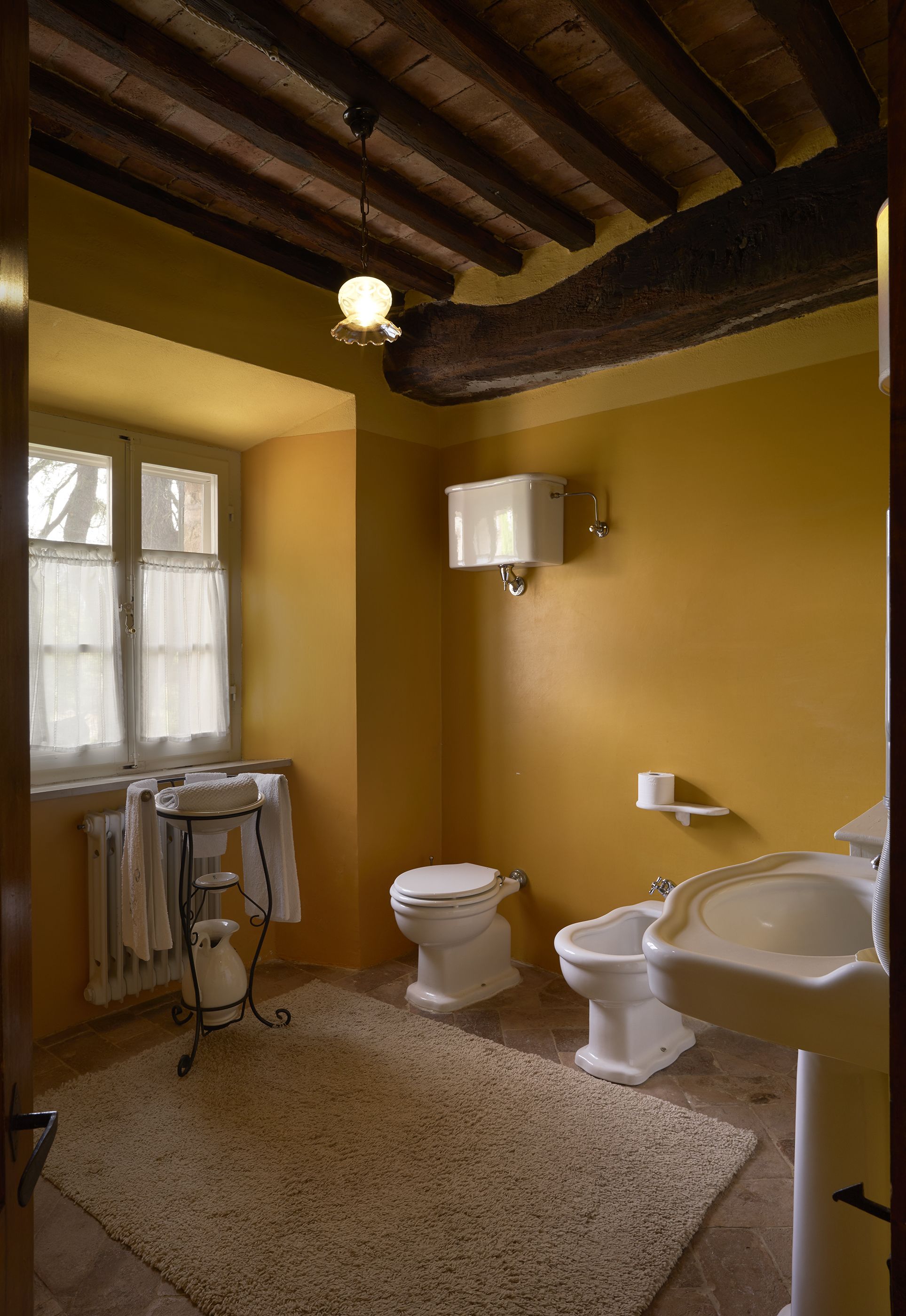 A bathroom with a toilet, bidet, and sink, wooden beams on the ceiling, and a window with curtains.