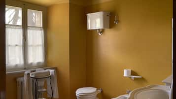 A bathroom with a toilet, bidet, and sink, wooden beams on the ceiling, and a window with curtains.